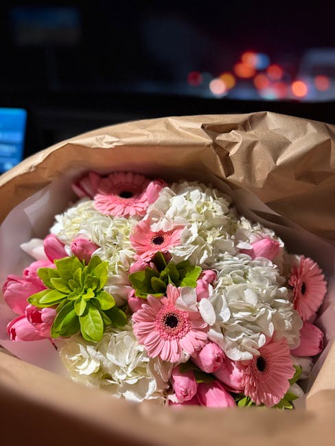 A flower bouquet featuring white hydrangeas and bright pink gerbera daisies, accented with fresh green foliage. The flowers are arranged in a natural, rounded shape, wrapped in brown craft paper with 