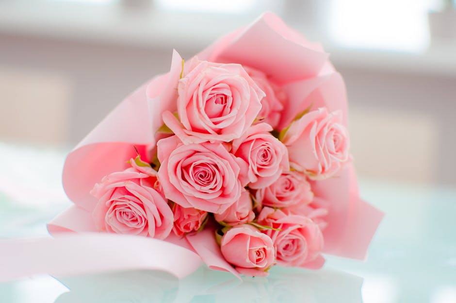 A bouquet of fresh pink roses arranged in a compact cluster, with tightly coiled petals and lush green stems visible beneath. The roses are wrapped in soft pink tissue paper, secured with a satin ribb