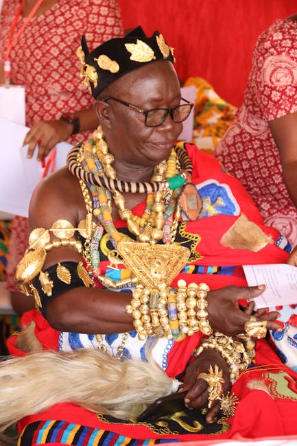 The image features an elderly woman wearing traditional African attire, including vibrant red fabric with intricate patterns and numerous gold and beaded jewelry, such as necklaces, bracelets, and ear
