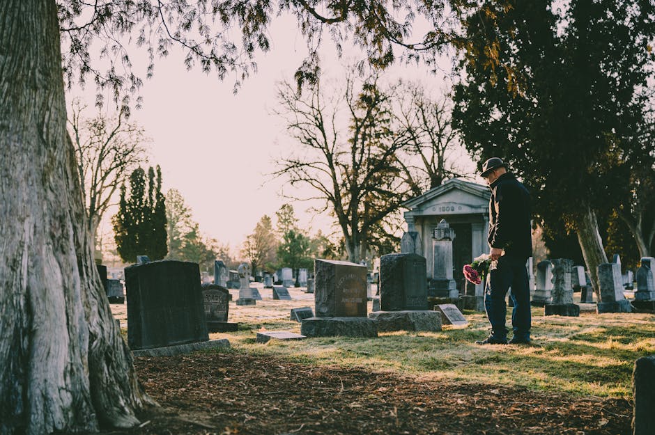 A man dressed in black formal attire and a hat stands in a cemetery during late afternoon or early evening, holding a bouquet of pink and red flowers with green foliage. Surrounding him are numerous u